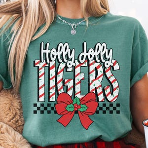 May include: Light green t-shirt with the text "Holly Jolly Tigers" in white and red candy cane stripes. A red bow with holly accents is below the text. The shirt is paired with red and black plaid pajama pants.