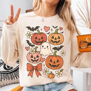 May include: Ivory-colored long-sleeve shirt with a Halloween-themed graphic featuring pumpkins, bats, flowers, a ghost, and a bow. An orange mug with a Halloween design is held in the hand. The word "ivory" is printed at the bottom.