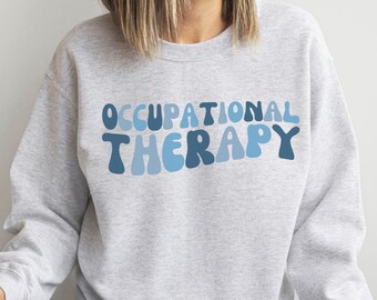 May include: A light gray sweatshirt with the words "OCCUPATIONAL THERAPY" in blue lettering. The text has a retro, wavy design. The sweatshirt is part of a clothing line.