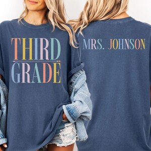 Op de afbeelding: Een blauw t-shirt met korte mouwen met de tekst THIRD GRADE in veelkleurige letters met een potloodafbeelding tussen de woorden op de voorkant, en de tekst MRS. JOHNSON in veelkleurige letters op de achterkant van het kledingstuk.