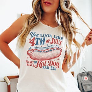 Puede incluir: Camiseta sin mangas color marfil con un gr&aacute;fico que dice "You look like the 4th of July makes me want a hot dog real bad." El gr&aacute;fico presenta una ilustraci&oacute;n de perrito caliente rojo, blanco y azul.