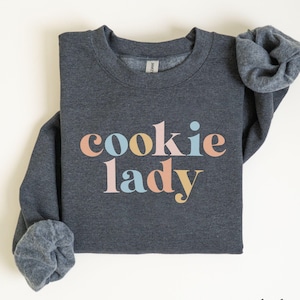 May include: Dark grey heather sweatshirt with the text "cookie lady" in colorful letters.
