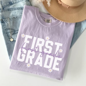 Op de afbeelding: Een lavendelkleurig T-shirt met de tekst "FIRST first grade" in wit met een paarse omtrek. De tekst wordt omringd door kleine witte madeliefjes met gele centra.