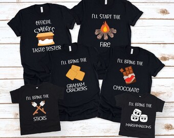 May include: Six black t-shirts with different designs for a camping trip. The shirts say "I'll start the fire", "I'll bring the graham crackers", "I'll bring the chocolate", "I'll bring the marshmallows", "I'll bring the sticks", and "Official Smores Taste Tester".