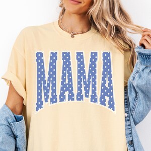 May include: Butter-colored t-shirt with the word "MAMA" in large, blue, polka-dotted letters across the front. The shirt has short sleeves and a relaxed fit. A light blue denim jacket is worn over the shirt.