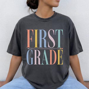 May include: A charcoal gray short-sleeve t-shirt featuring the text FIRST GRADE in a multi-colored serif font. The word FIRST is stacked above the word GRADE, separated by a horizontal line graphic resembling a pencil with a pointed tip.