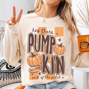 May include: An ivory long-sleeve sweatshirt featuring a graphic print with the text "hey there PUMPKIN pick of the patch" and "100% FRESH". The design includes orange checkered blocks, two orange pumpkins, and small star accents. The sweatshirt has a crew neckline.