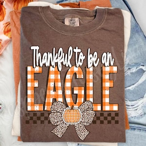 May include: A brown short-sleeve t-shirt featuring the white text Thankful to be an EAGLE. The word EAGLE is in an orange and white gingham pattern. Below the text is a leopard print bow with a small orange and white gingham pumpkin in the center.
