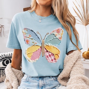 May include: Light blue t-shirt featuring a large butterfly applique. The butterfly design incorporates various fabric patterns, including floral, gingham, and solid colors. The word "chambray" is printed at the bottom left of the image. The shirt has short sleeves.