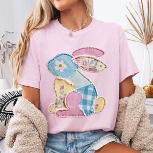 May include: A light pink t-shirt featuring a patchwork bunny design. The bunny is composed of various fabric patterns in blue, yellow, pink, and floral prints. The word "blossom" is printed in the bottom left corner.