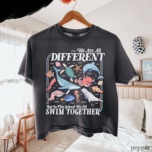 May include: Dark gray t-shirt featuring a colorful graphic of various sea creatures, including a whale, octopus, and dolphin. The text reads "We Are All DIFFERENT But In This School We All SWIM TOGETHER."