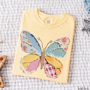 May include: A yellow Comfort Colors t-shirt with a large butterfly applique. The butterfly wings are made of patchwork fabric in shades of blue, pink, yellow, and red. The shirt is folded and lying on a white textured surface. The word "butter" is visible.