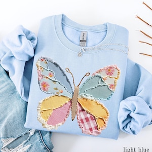 May include: Light blue sweatshirt with a patchwork butterfly design. The butterfly features various fabric patterns in shades of blue, yellow, pink, and red. A silver necklace with a small pendant is visible. The text "light blue" is at the bottom right.