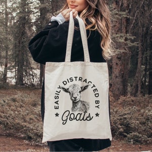 May include: A white canvas tote bag with black text that reads "Easily Distracted By Goats" in a circle with a goat's head in the center. The tote bag has black straps.