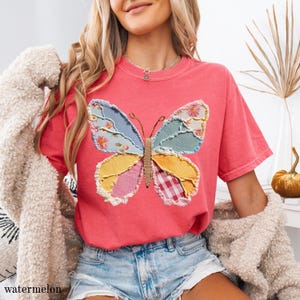 May include: A watermelon-colored t-shirt features a patchwork butterfly design. The butterfly's wings are composed of various fabric patterns, including floral, gingham, and solid colors. The shirt has a relaxed fit and short sleeves. The word "watermelon" is printed at the bottom left.