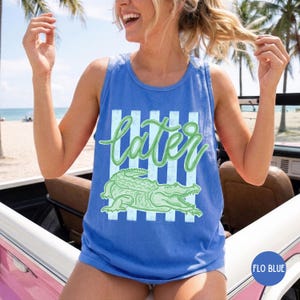 May include: A blue tank top with the word "later" in green script, above a green alligator illustration. The design is set against a backdrop of vertical light blue stripes. The tank top is a "Flo Blue" color.
