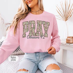 May include: Light pink sweatshirt with the word "PRAY" in a camouflage pattern across the chest. The text "Colossians 4:2" is below the word "PRAY". The sweatshirt is long-sleeved and made of a soft material.