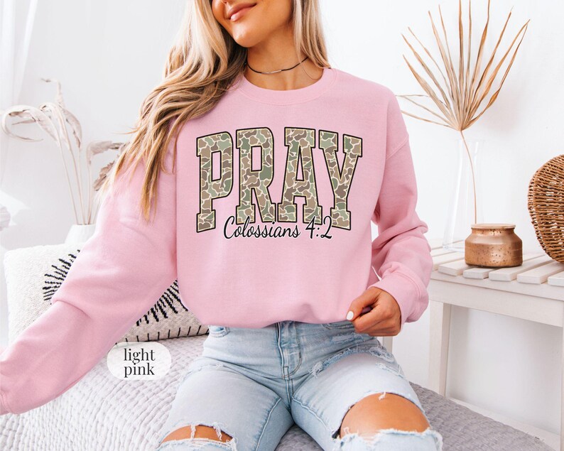 May include: Light pink sweatshirt with the word "PRAY" in a camouflage pattern across the chest. The text "Colossians 4:2" is below the word "PRAY". The sweatshirt is long-sleeved and made of a soft material.