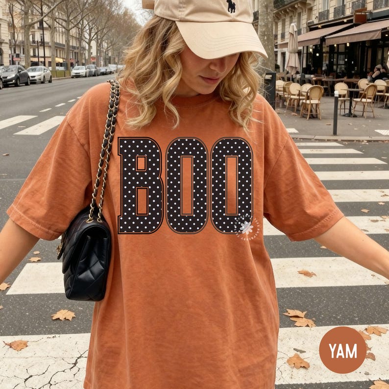 Puede incluir: Una camiseta oversize de color naranja quemado con la palabra BOO impresa en grandes letras negras con un patr&oacute;n de lunares blancos. Se combina con un bolso de hombro negro acolchado con cadena dorada y una gorra de b&eacute;isbol de color beige.