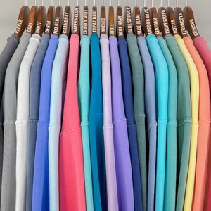 May include: A display of sweatshirts in a spectrum of colours, from grey and white to pink and green, suspended on wooden hangers. Each hanger is labelled with the sweatshirt's colour name, including "Pepper", "Denim", and "Crimson".