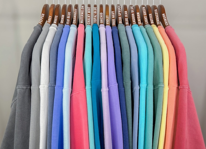 May include: A display of sweatshirts in a spectrum of colours, from grey and white to pink and green, suspended on wooden hangers. Each hanger is labelled with the sweatshirt's colour name, including "Pepper", "Denim", and "Crimson".