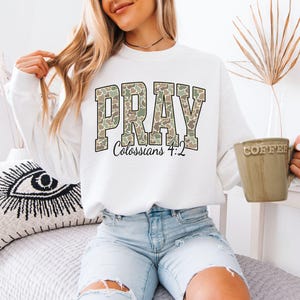 May include: White sweatshirt with the word "PRAY" in a camouflage pattern, with "Colossians 4:2" below. The sweatshirt is paired with light blue denim shorts. A beige coffee mug is held in the hand.