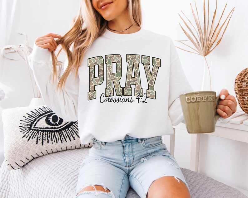 May include: White sweatshirt with the word "PRAY" in a camouflage pattern, with "Colossians 4:2" below. The sweatshirt is paired with light blue denim shorts. A beige coffee mug is held in the hand.