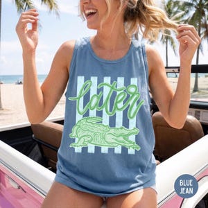 May include: Blue jean tank top with a green alligator graphic and the word "later" in a cursive font. The tank top has a relaxed fit and is made of a soft material. The design features vertical stripes behind the text and alligator.