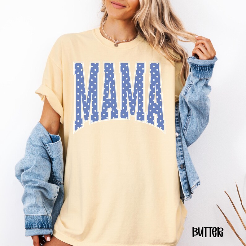May include: Butter-colored t-shirt with the word "MAMA" in large, blue, polka-dotted letters across the front. The shirt has short sleeves and a relaxed fit. A light blue denim jacket is worn over the shirt.