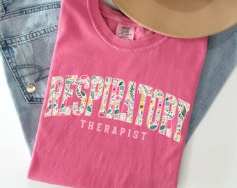 May include: Pink t-shirt with a floral design that spells out "Respiratory Therapist".