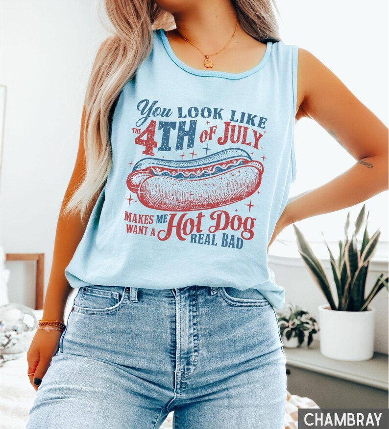 Puede incluir: Camiseta sin mangas azul claro con un gr&aacute;fico que dice "You look like the 4th of July makes me want a hot dog real bad." El gr&aacute;fico presenta una ilustraci&oacute;n de perrito caliente rojo y azul. La camiseta sin mangas es sin mangas.