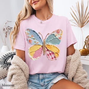 May include: A light pink t-shirt featuring a colorful butterfly applique. The butterfly design incorporates various fabric patterns in shades of blue, yellow, pink, and red. The word "blossom" is printed in the lower left corner.