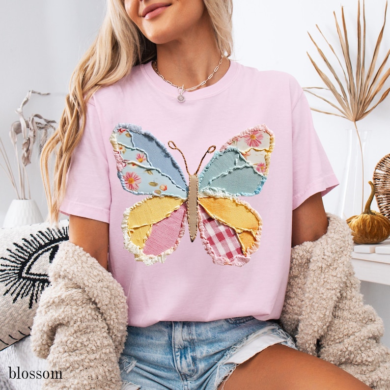 May include: A light pink t-shirt featuring a colorful butterfly applique. The butterfly design incorporates various fabric patterns in shades of blue, yellow, pink, and red. The word "blossom" is printed in the lower left corner.