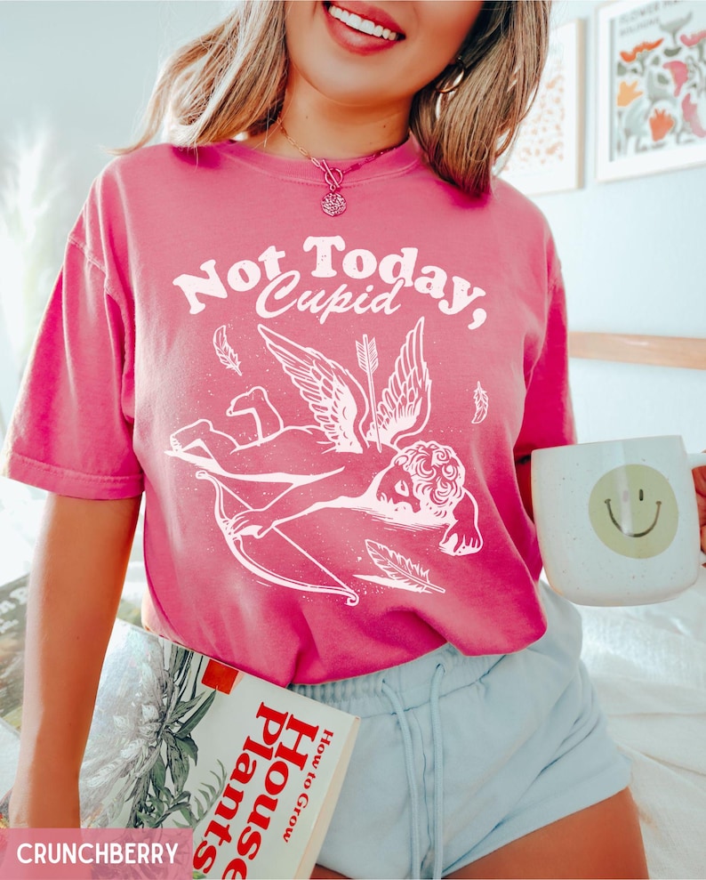 May include: Pink t-shirt with a white graphic of a winged cupid with an arrow and the text "Not Today, Cupid".