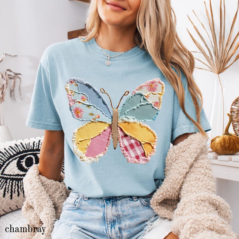 May include: Light blue t-shirt featuring a large butterfly applique. The butterfly design incorporates various fabric patterns, including floral, gingham, and solid colors. The word "chambray" is printed at the bottom left of the image. The shirt has short sleeves.