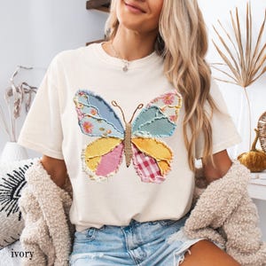 May include: Ivory-colored t-shirt featuring a colorful patchwork butterfly design. The butterfly has wings in shades of blue, yellow, pink, and red, with floral and gingham patterns. The shirt is a casual style.