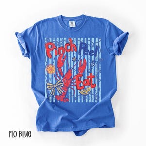 Op de afbeelding: Een blauw t-shirt met een grafisch ontwerp met een rode rivierkreeft, de woorden "Pinch Peel Eat" en decoratieve elementen. Het shirt is effen blauw van kleur met opgerolde mouwen.