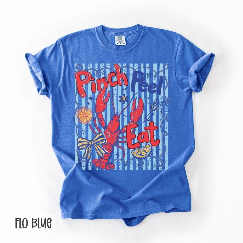 Op de afbeelding: Een blauw t-shirt met een grafisch ontwerp met een rode rivierkreeft, de woorden "Pinch Peel Eat" en decoratieve elementen. Het shirt is effen blauw van kleur met opgerolde mouwen.