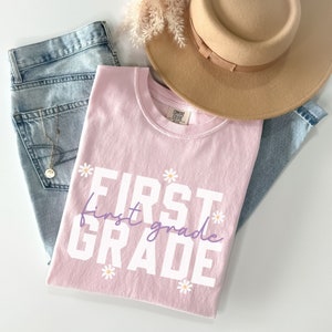Op de afbeelding: Roze T-shirt met de tekst "FIRST grade" in witte letters met een paarse "first grade" tekst eronder. Het shirt is versierd met kleine witte madeliefjes.