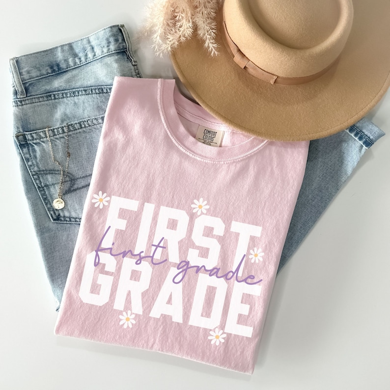 Op de afbeelding: Roze T-shirt met de tekst "FIRST grade" in witte letters met een paarse "first grade" tekst eronder. Het shirt is versierd met kleine witte madeliefjes.