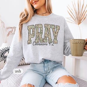 May include: A light gray sweatshirt with the word "PRAY" in a camouflage pattern, with "Colossians 4:2" below. The sweatshirt is ash-colored and has a crew neck. A green coffee mug is held in the hand.