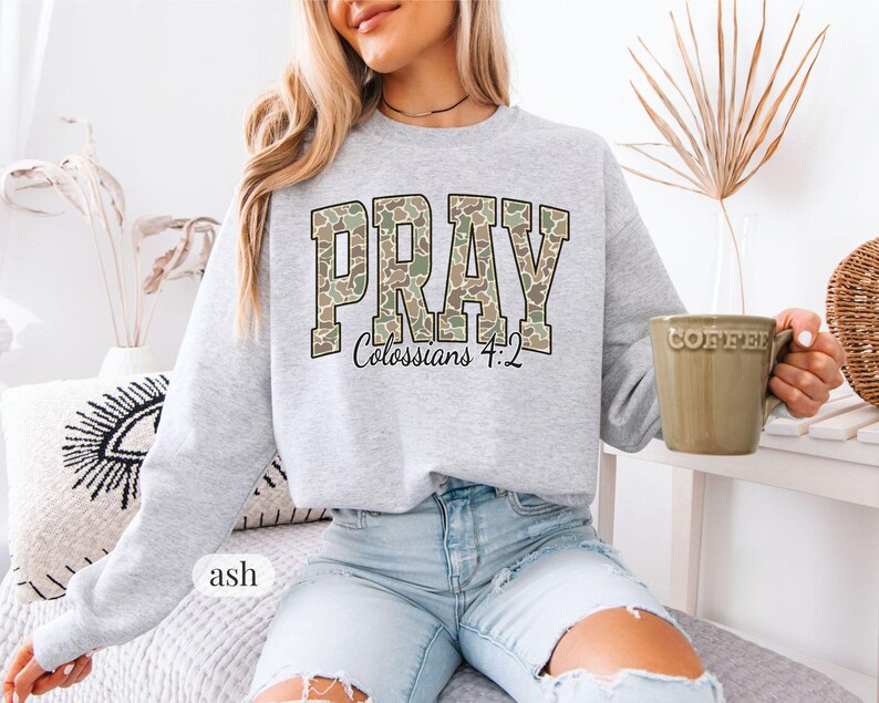 May include: A light gray sweatshirt with the word "PRAY" in a camouflage pattern, with "Colossians 4:2" below. The sweatshirt is ash-colored and has a crew neck. A green coffee mug is held in the hand.