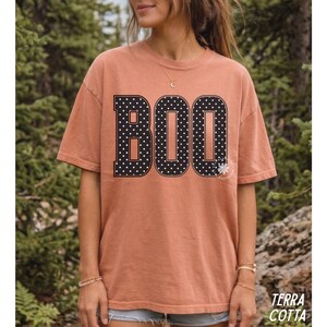 Puede incluir: Una camiseta de manga corta color terracota con la palabra BOO impresa en letras may&uacute;sculas grandes, negritas y negras. Cada letra est&aacute; rellena con un patr&oacute;n de lunares blancos. La camiseta tiene cuello redondo y un corte holgado.