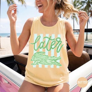 May include: Yellow tank top with the word "later" in green script, a green alligator graphic, and light blue vertical stripes. The tank top is a casual, sleeveless style.
