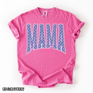 May include: A pink t-shirt with the word "MAMA" in large, blue letters with white polka dots. The shirt has rolled-up sleeves and the word "CRUNCHBERRY" is printed at the bottom.