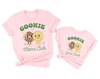 May include: Two pink t-shirts with a cookie and lemon graphic. The larger shirt says "Cookie Moms Club" and the smaller shirt says "Cookie Girls Club".