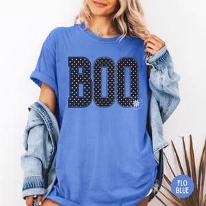 Puede incluir: Una camiseta de manga corta color azul aciano con la palabra BOO impresa en letras grandes de estilo bloque. Las letras son negras con un patr&oacute;n de lunares blancos y un borde blanco fino. Un peque&ntilde;o icono de margarita blanca aparece junto a la O.