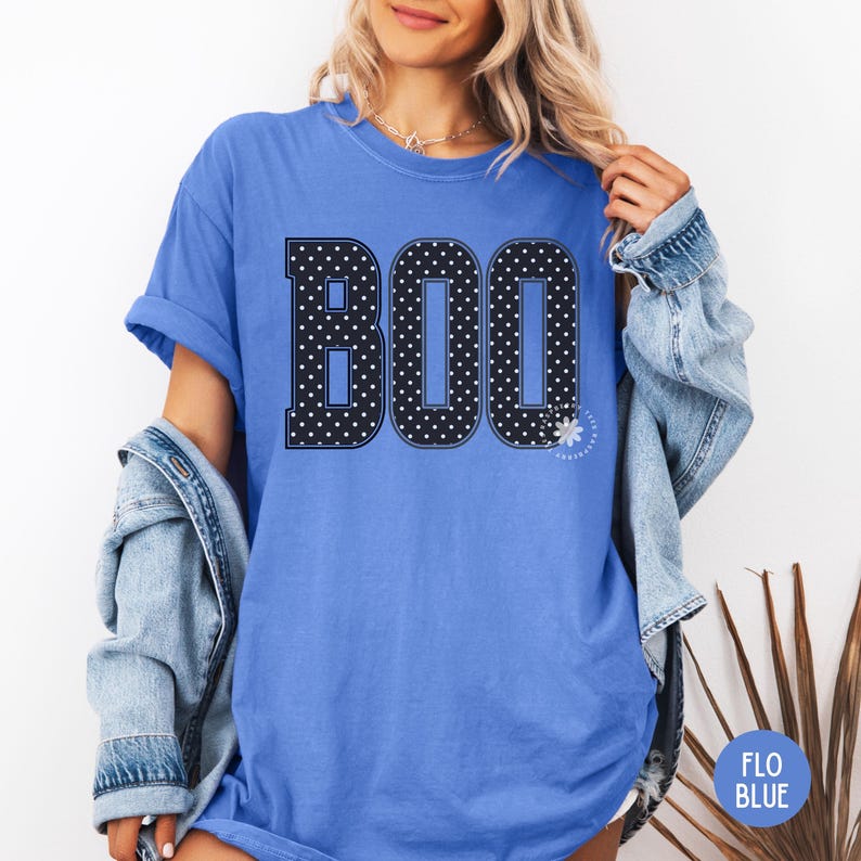 Puede incluir: Una camiseta de manga corta color azul aciano con la palabra BOO impresa en letras grandes de estilo bloque. Las letras son negras con un patr&oacute;n de lunares blancos y un borde blanco fino. Un peque&ntilde;o icono de margarita blanca aparece junto a la O.