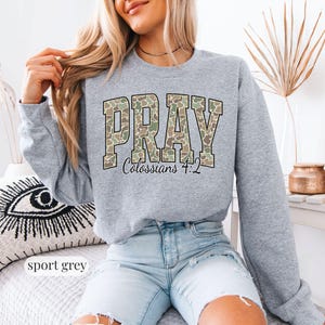 May include: Sport grey sweatshirt with the word "PRAY" in a camouflage pattern across the chest. The text "Colossians 4:2" is below the word "PRAY". The sweatshirt is a crew neck style.