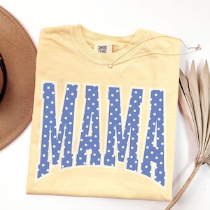 May include: A light yellow t-shirt with the word "MAMA" in large blue letters with white polka dots. The shirt has a crew neck and short sleeves. A brown hat and a gold necklace are also in the image.